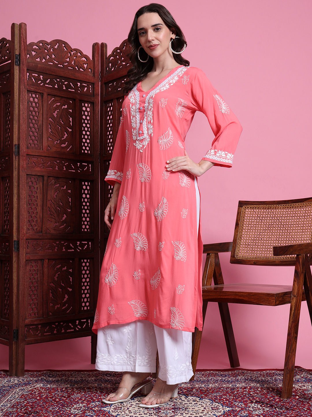 Woman wearing a pink kurta with white patterns in a room with wooden furniture and a pink wall.