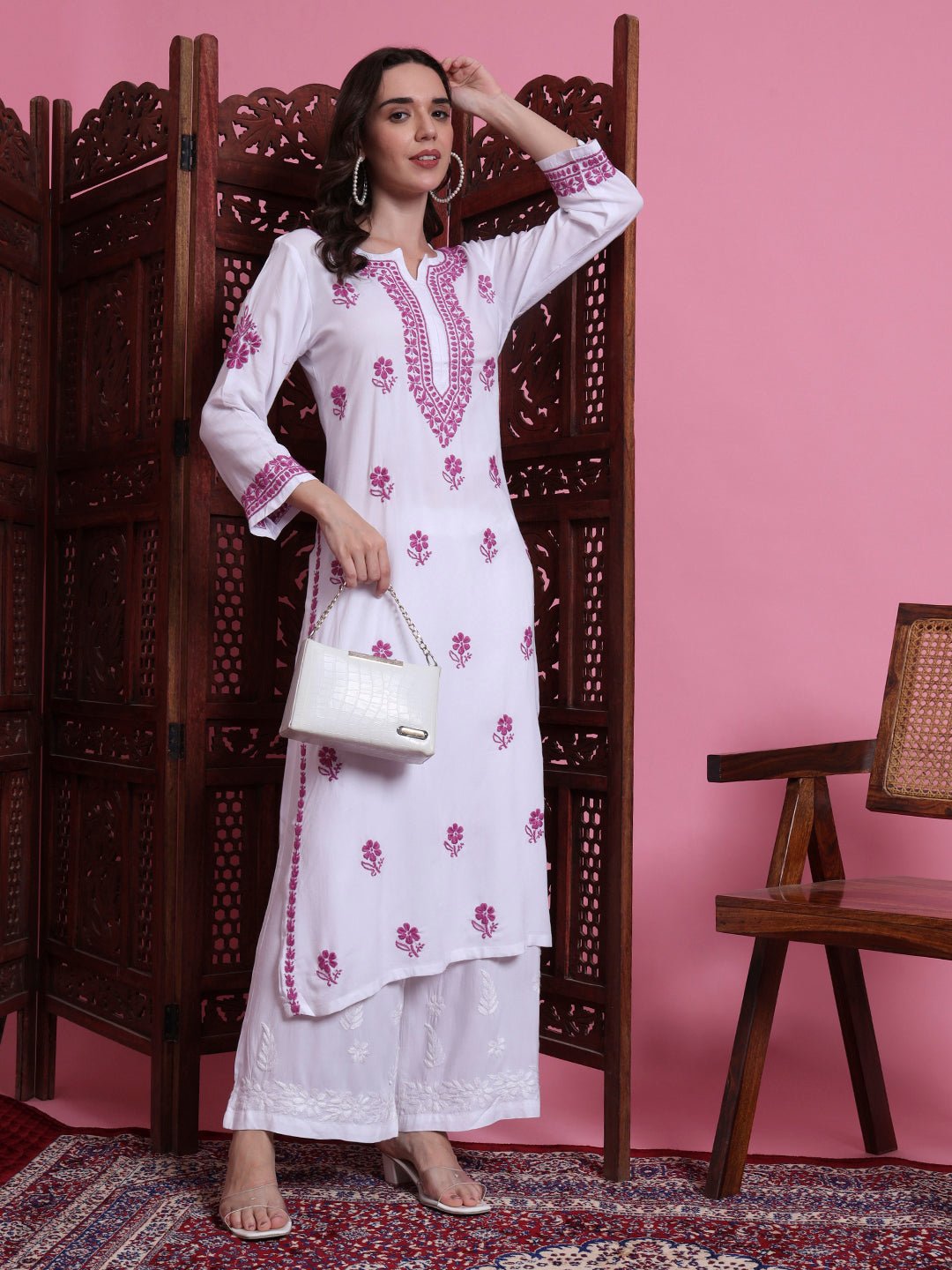 Woman in a white embroidered outfit with pink floral patterns against a pink wall.
