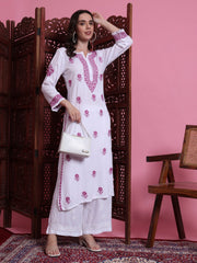 Woman in a white embroidered outfit with pink floral patterns against a pink wall.
