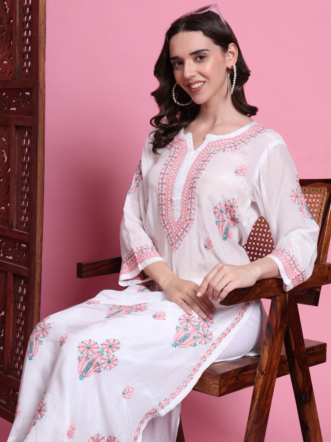 Woman wearing a white embroidered outfit sitting on a wooden chair against a pink background