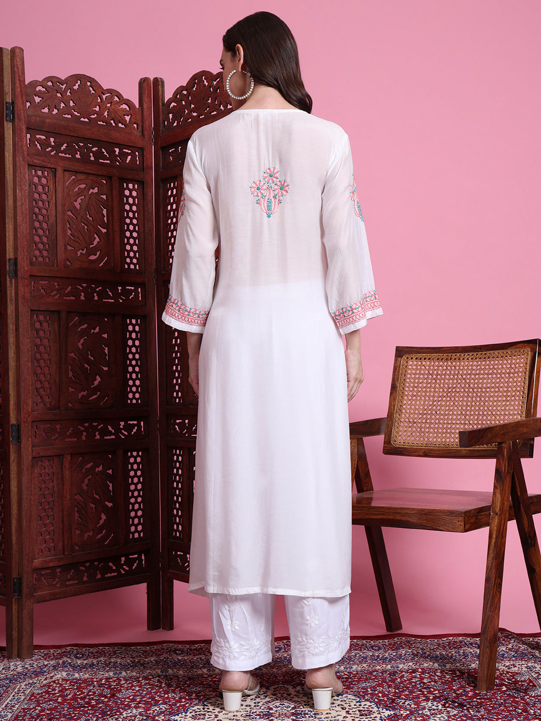 Woman wearing a white kurta with pink embroidery in a room with pink walls and wooden furniture.