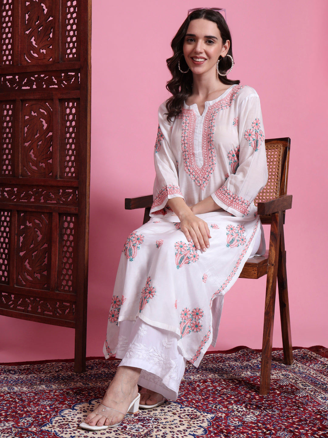 Woman in a white embroidered outfit sitting on a wooden chair against a pink wall.
