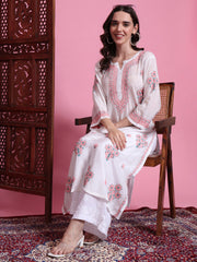 Woman in a white embroidered outfit sitting on a wooden chair against a pink wall.